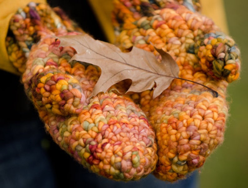 May include: A pair of colorful crocheted mittens with a brown leaf resting on top. The mittens are made with a variety of shades of orange, yellow, pink, and red yarn.