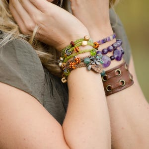 May include: A collection of layered bracelets featuring green and brown braided cords with orange, green, and wooden beads, a purple beaded bracelet with faceted stones, and a wide brown leather cuff bracelet with metal eyelets and lace-up details.