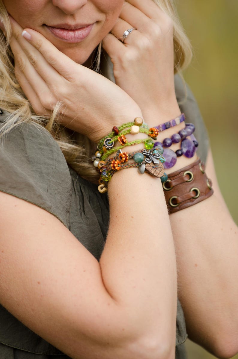 May include: A collection of layered bracelets featuring green and brown braided cords with orange, green, and wooden beads, a purple beaded bracelet with faceted stones, and a wide brown leather cuff bracelet with metal eyelets and lace-up details.
