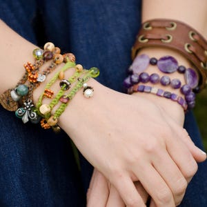 May include: A collection of layered bracelets featuring green braided cord with assorted wooden, glass, and pearl beads, alongside a brown leather cuff with metal eyelets and a stack of purple gemstone bead bracelets with a square-cut pattern.