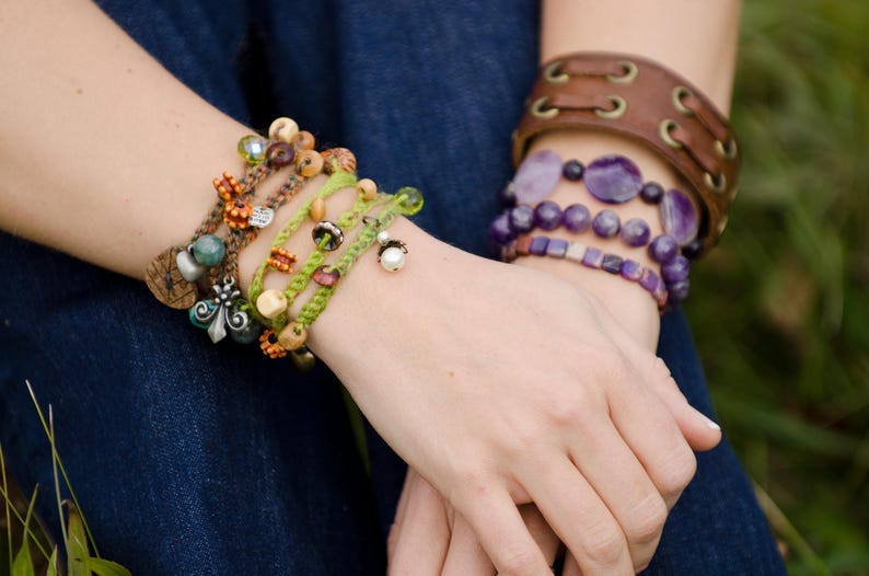 May include: A collection of layered bracelets featuring green braided cord with assorted wooden, glass, and pearl beads, alongside a brown leather cuff with metal eyelets and a stack of purple gemstone bead bracelets with a square-cut pattern.