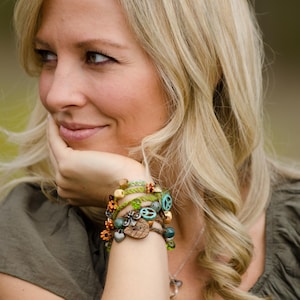 May include: A multi-strand wrap bracelet featuring green braided cord, wooden beads, metal charms including a peace sign, heart, and flower shapes, and small turquoise-coloured beads, worn around the wrist as a layered accessory piece.