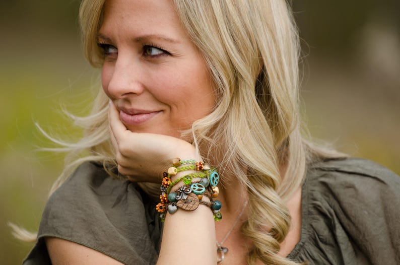 May include: A multi-strand wrap bracelet featuring green braided cord, wooden beads, metal charms including a peace sign, heart, and flower shapes, and small turquoise-colored beads, worn around the wrist as a layered accessory piece.