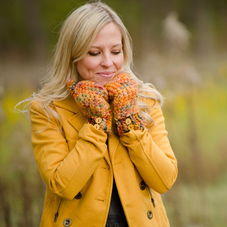 May include: A woman wearing a yellow coat with a pair of colorful crocheted fingerless gloves. The gloves have a floral button detail.