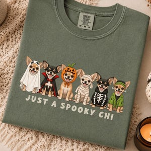 May include: A moss green crewneck t-shirt featuring a graphic of six Chihuahua dogs in various Halloween costumes, including a ghost, vampire, pumpkin, mummy, skeleton, and Frankenstein. The text below reads, "JUST A SPOOKY CHI."