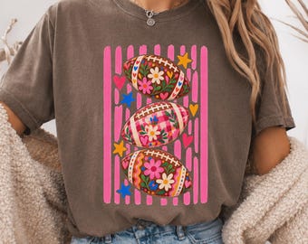 Puede incluir: Una camiseta de manga corta color espresso con un estampado gráfico de tres balones de fútbol americano apilados verticalmente. Cada balón está decorado con coloridos motivos florales y cuadros vichy, rodeado de rayas verticales rosas.