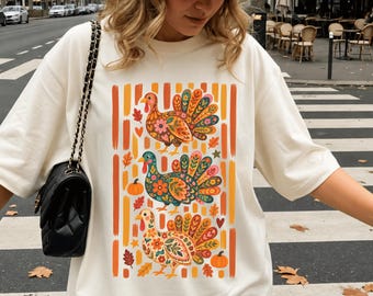 Puede incluir: Una camiseta color marfil con un estampado gráfico de tres pavos con coloridos patrones florales, rodeados de rayas verticales naranjas, pequeñas calabazas y hojas de otoño, acompañada de una gorra de béisbol beige y un bolso de hombro negro.