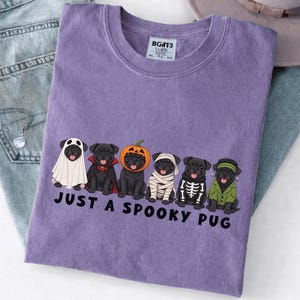 Könnte beinhalten: Ein violettes T-Shirt mit einer Grafik von sechs schwarzen Möpsen in verschiedenen Halloween-Kostümen, darunter ein Geist, Vampir, Kürbis, Mumie, Skelett und Frankensteins Monster, mit dem schwarzen Schriftzug "JUST A SPOOKY PUG" darunter.
