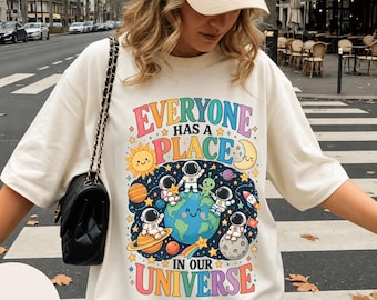 Puede incluir: Una camiseta de manga corta color marfil con un estampado gráfico colorido de astronautas, planetas, estrellas y una Tierra sonriente. El texto dice "EVERYONE HAS A PLACE IN OUR UNIVERSE" en letras multicolores. Se lleva con una gorra beige.