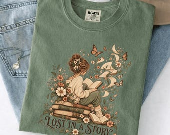 Puede incluir: Una camiseta verde salvia con un gráfico de una persona sentada sobre una pila de libros leyendo, rodeada de flores, mariposas y páginas flotantes. El texto "LOST IN A STORY" está impreso en marrón debajo de la ilustración.