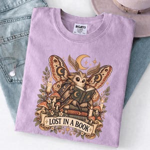 May include: A light purple t-shirt featuring a graphic of a moth reading a book while sitting on a stack of books, surrounded by mushrooms, candles, and foliage. The text "LOST IN A BOOK" is printed on a banner at the bottom of the illustration.