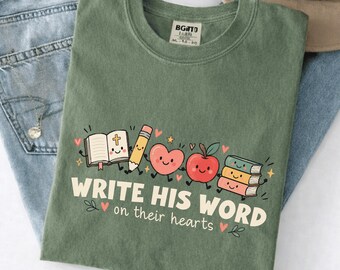 Puede incluir: Una camiseta verde musgo con un gráfico de una Biblia abierta sonriente, un lápiz, un corazón, una manzana y una pila de libros. El texto "WRITE HIS WORD on their hearts" aparece en letras color crema, decorado con pequeños corazones y estrellas.