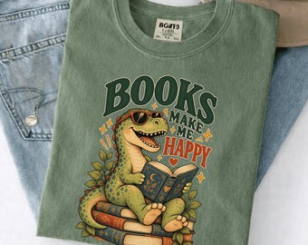 Puede incluir: Una camiseta color verde musgo con el gráfico de un dinosaurio verde con gafas de sol leyendo un libro abierto sobre una pila de libros. El texto "BOOKS MAKE ME HAPPY" aparece en letras de color verde oscuro y rojo con detalles de estrellas y corazones.