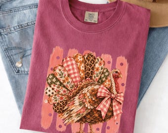 Puede incluir: Una camiseta color baya con el gráfico de un pavo cuya cola está formada por plumas con diversos patrones, incluyendo flores, cuadros vichy y estampado de leopardo. El pavo lleva un lazo de cuadros vichy rojo y blanco. Cuello redondo.