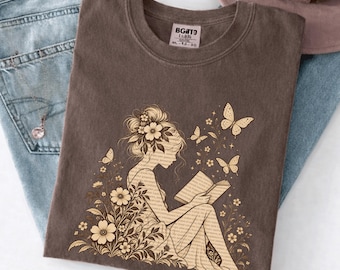 Puede incluir: Una camiseta de color marrón oscuro con un gráfico de una persona con flores en el pelo leyendo un libro, rodeada de mariposas y elementos florales. El gráfico está en un estilo de línea color canela claro. La etiqueta dice BGTD.