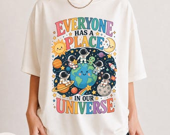 Puede incluir: Una camiseta de color marfil con un gráfico colorido del espacio, planetas, estrellas, una Tierra sonriente y astronautas con el texto "EVERYONE HAS A PLACE IN OUR UNIVERSE" en una fuente divertida. El diseño incluye un sol, una luna y un cohete.