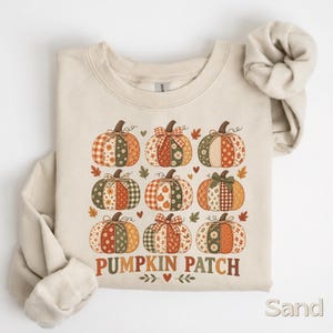 May include: A sand-coloured crewneck sweatshirt featuring a graphic print of nine pumpkins arranged in a grid. Each pumpkin displays a different patchwork pattern in shades of orange, green, and brown. The text PUMPKIN PATCH is printed below.