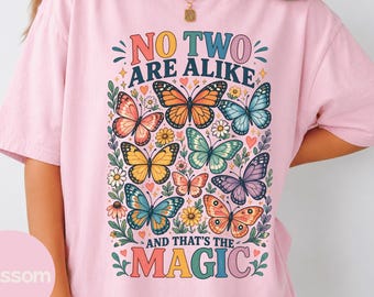 Puede incluir: Una camiseta de manga corta de color rosa claro con un gráfico colorido de varias mariposas y flores. El texto dice NO TWO ARE ALIKE AND THAT'S THE MAGIC. El diseño incluye ilustraciones de mariposas naranjas, azules, amarillas, verdes y moradas.
