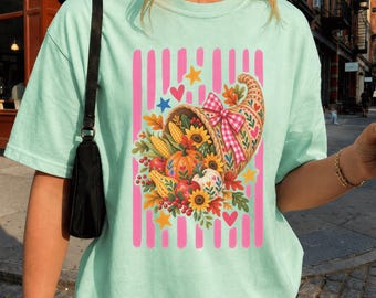 Puede incluir: Una camiseta de color verde menta claro con un gráfico central de un cuerno de la abundancia lleno de productos de cosecha otoñal como calabazas, girasoles y maíz. El diseño incluye un lazo de cuadros rosa, rayas verticales rosas y estrellas.