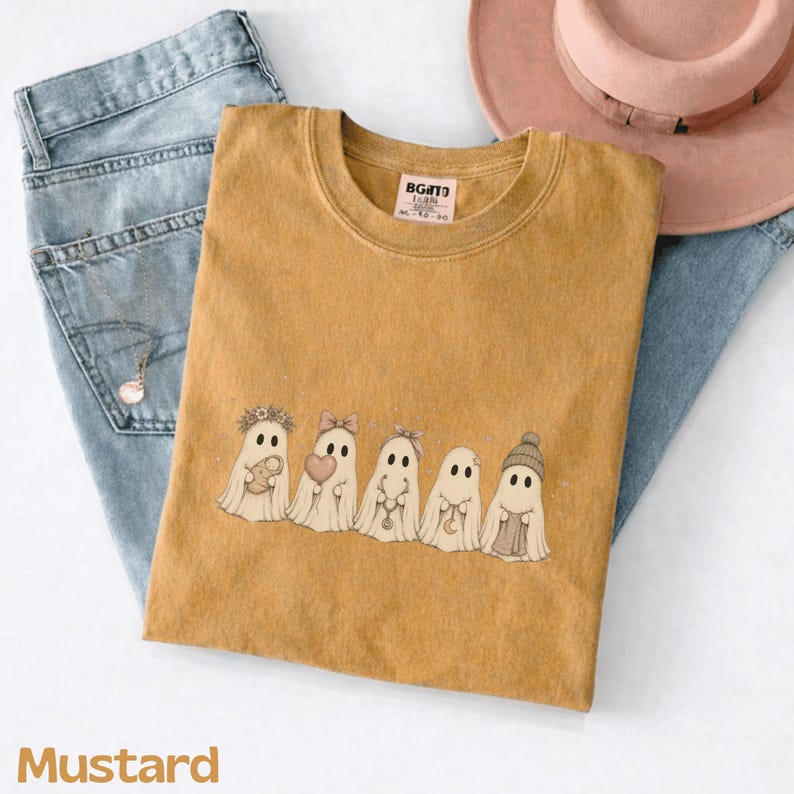 May include: A mustard yellow t-shirt featuring a graphic of five white ghosts. The ghosts are wearing accessories including a flower crown, a bow, a headband, a crescent moon necklace, and a beanie. The shirt has a tag reading BGITD M-10-20.