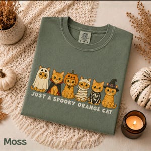 May include: A moss green crewneck t-shirt featuring a graphic of six orange cats in various Halloween costumes, including a ghost, vampire, pumpkin, mummy, skeleton, and witch. The text below the cats reads, "JUST A SPOOKY ORANGE CAT."