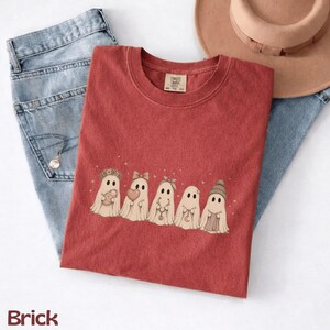 May include: A brick red crew neck t-shirt featuring a graphic of five ghosts in a row. The ghosts are decorated with a flower crown, a bow, a headband, a crescent moon necklace, and a beanie. The shirt has a tag reading Comfort Colors.