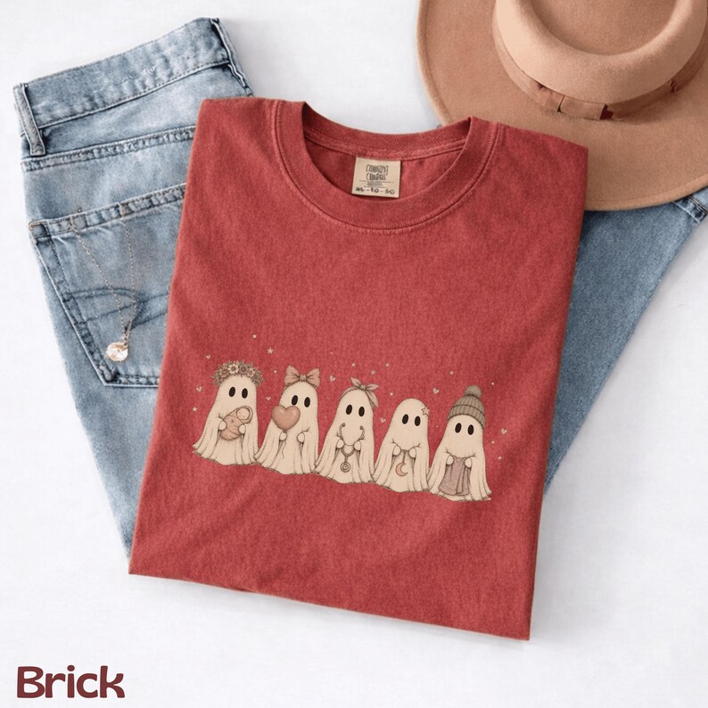May include: A brick red crew neck t-shirt featuring a graphic of five ghosts in a row. The ghosts are decorated with a flower crown, a bow, a headband, a crescent moon necklace, and a beanie. The shirt has a tag reading Comfort Colors.