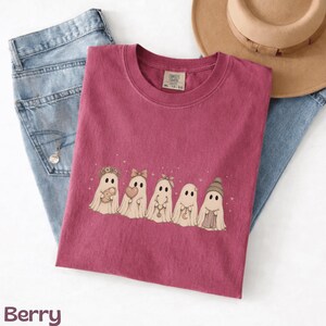 May include: A berry-coloured crewneck t-shirt featuring a graphic of five ghosts in a row. The ghosts are decorated with accessories including a flower crown, a bow, a headband, a necklace, and a beanie. The shirt has a tag reading Comfort Colors.
