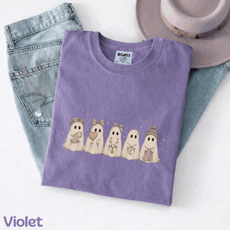 May include: A violet crew neck t-shirt featuring a graphic of five beige ghosts in a row. The ghosts are decorated with accessories including a flower crown, a bow, a necklace, a crescent moon charm, and a knit beanie. The shirt has a white tag.