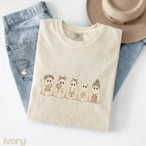 May include: An ivory crew neck t-shirt featuring a graphic print of five cartoon ghosts in a row. The ghosts are decorated with accessories including a flower crown, a bow, a headband, and a knit beanie, while holding items like a heart and a moon.