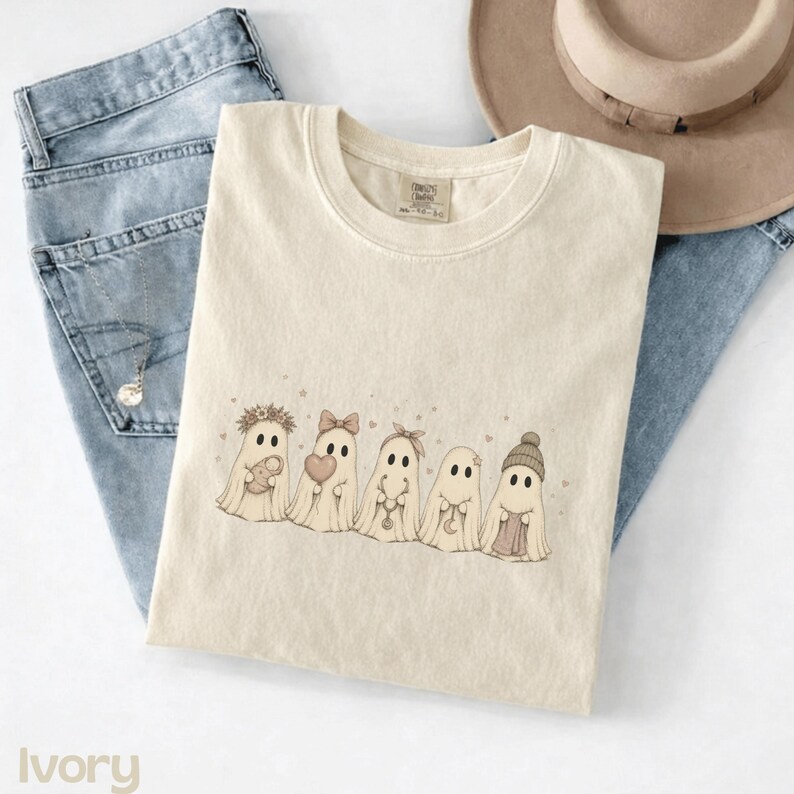 May include: An ivory crew neck t-shirt featuring a graphic print of five cartoon ghosts in a row. The ghosts are decorated with accessories including a flower crown, a bow, a headband, and a knit beanie, while holding items like a heart and a moon.