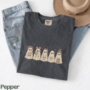 May include: A charcoal grey t-shirt featuring a graphic of five cartoon ghosts in a row. The ghosts are decorated with accessories including a flower crown, a bow, a headband, a necklace, and a beanie, with small stars and hearts surrounding them.