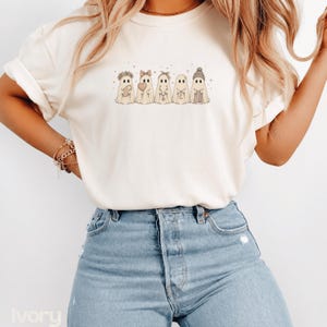 May include: An ivory short-sleeved t-shirt featuring a graphic print of five ghosts in a row. The ghosts are decorated with various accessories including a flower crown, a bow, a headband, a necklace, and a beanie, accented by small stars.