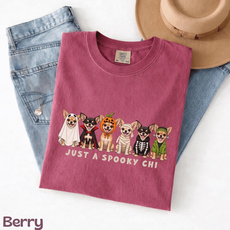 May include: A berry-colored t-shirt featuring a graphic of six Chihuahua dogs in various Halloween costumes, including a ghost, vampire, pumpkin, mummy, skeleton, and Frankenstein. The text below the dogs reads, "JUST A SPOOKY CHI."