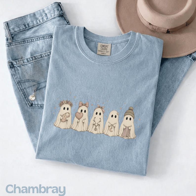 May include: A light blue t-shirt featuring a graphic of five ghosts in a row. The ghosts are wearing accessories including a flower crown, a bow, a beanie, and holding items like a small bundle, a heart, and a crescent moon charm. The shirt has a Comfort Colors label.