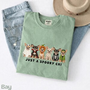 May include: A sage green t-shirt featuring a graphic of six Chihuahua dogs in various Halloween costumes, including a ghost, vampire, pumpkin, mummy, skeleton, and Frankenstein's monster, with the text "JUST A SPOOKY CHI" printed below.