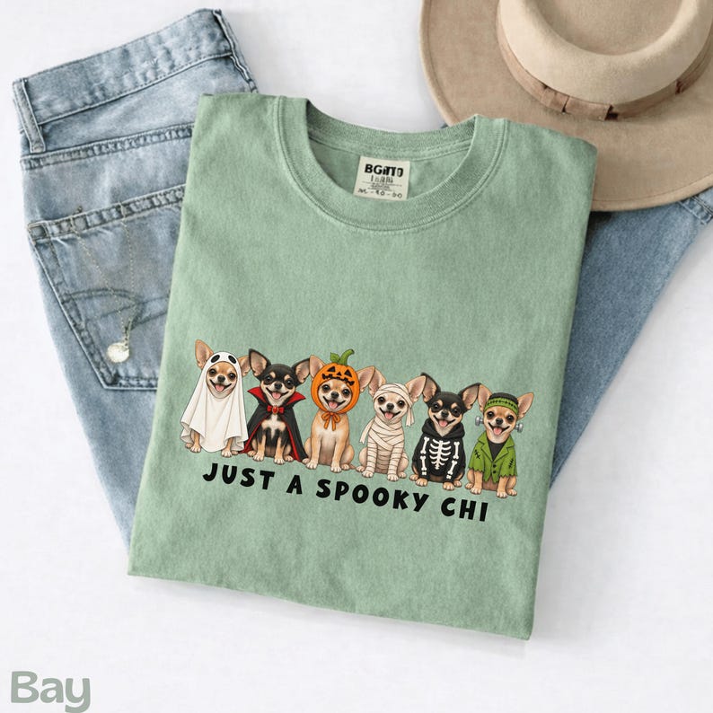 May include: A sage green t-shirt featuring a graphic of six Chihuahua dogs in various Halloween costumes, including a ghost, vampire, pumpkin, mummy, skeleton, and Frankenstein's monster, with the text "JUST A SPOOKY CHI" printed below.
