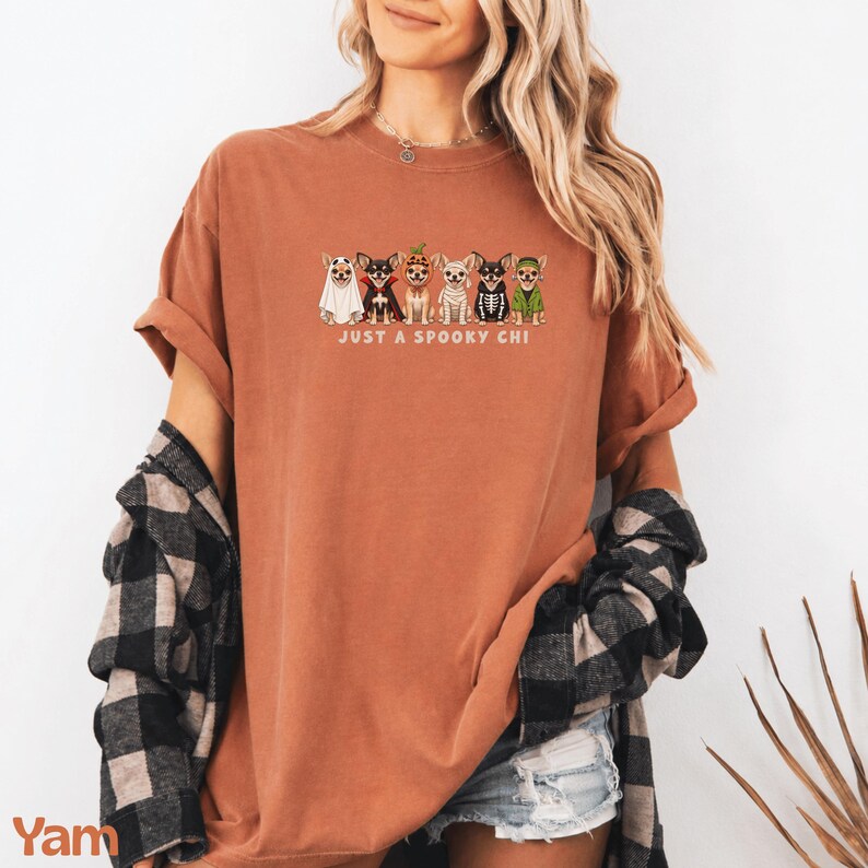May include: A rust-colored short-sleeve t-shirt featuring a graphic of six Chihuahua dogs in various Halloween costumes, including a ghost, vampire, pumpkin, mummy, skeleton, and witch, with the text "JUST A SPOOKY CHI" printed below the dogs.