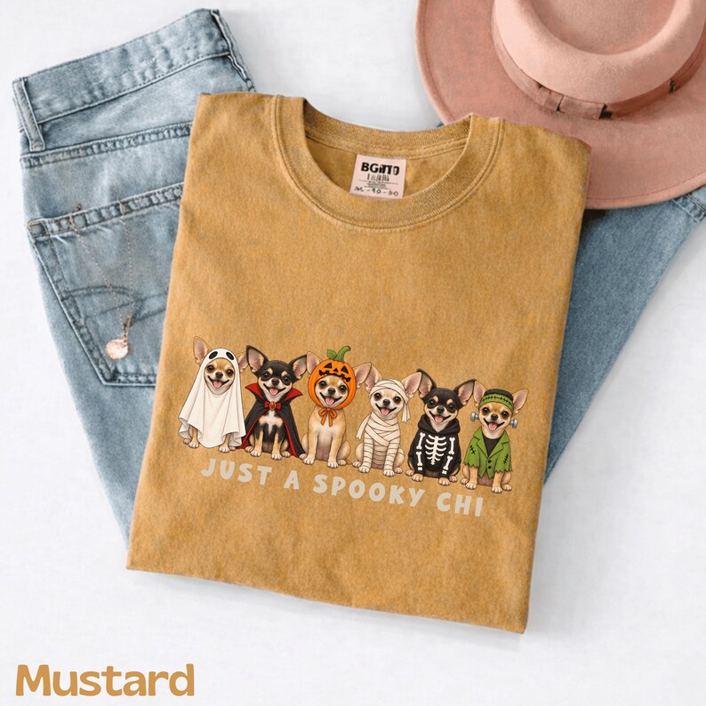May include: A mustard yellow t-shirt featuring a graphic of six Chihuahuas in various Halloween costumes, including a ghost, vampire, pumpkin, mummy, skeleton, and Frankenstein's monster, with the text "JUST A SPOOKY CHI" printed below.