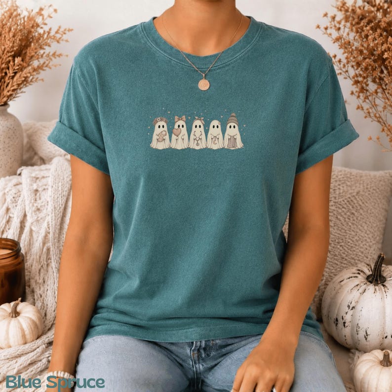 May include: A teal short-sleeve t-shirt featuring a graphic print of five white ghosts in a row. Each ghost has a different accessory, including a flower crown, a bow, a headband, a necklace, and a beanie. The shirt has a crew neckline.