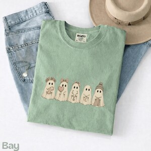 May include: A sage green t-shirt featuring a graphic of five cartoon ghosts in a row. The ghosts are decorated with accessories including a flower crown, a bow, a small heart, a crescent moon charm, and a knit beanie hat. The shirt has a crew neck.