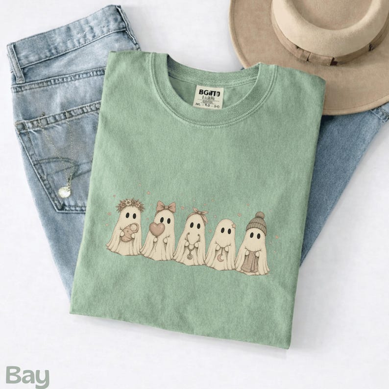 May include: A sage green t-shirt featuring a graphic of five cartoon ghosts in a row. The ghosts are decorated with accessories including a flower crown, a bow, a small heart, a crescent moon charm, and a knit beanie hat. The shirt has a crew neck.