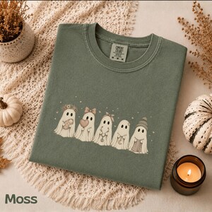 May include: A moss green crewneck t-shirt featuring a graphic of five white ghosts in a row. The ghosts are decorated with accessories including a flower crown, a bow, a headband, a necklace, and a beanie. The shirt has a Comfort Colors label.