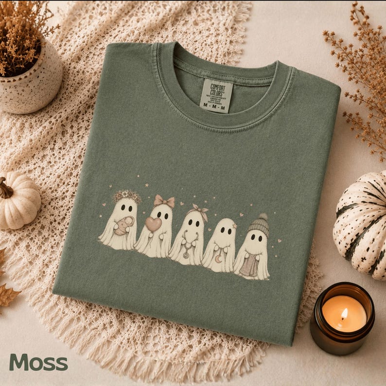 May include: A moss green crewneck t-shirt featuring a graphic of five white ghosts in a row. The ghosts are decorated with accessories including a flower crown, a bow, a headband, a necklace, and a beanie. The shirt has a Comfort Colors label.