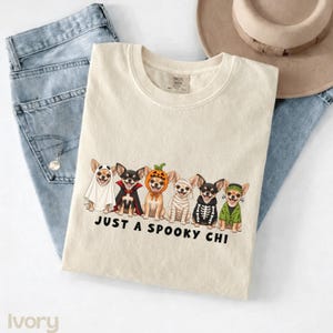 May include: An ivory crewneck t-shirt featuring a graphic of six Chihuahua dogs in various Halloween costumes, including a ghost, vampire, pumpkin, mummy, skeleton, and Frankenstein's monster, with the text JUST A SPOOKY CHI printed below.