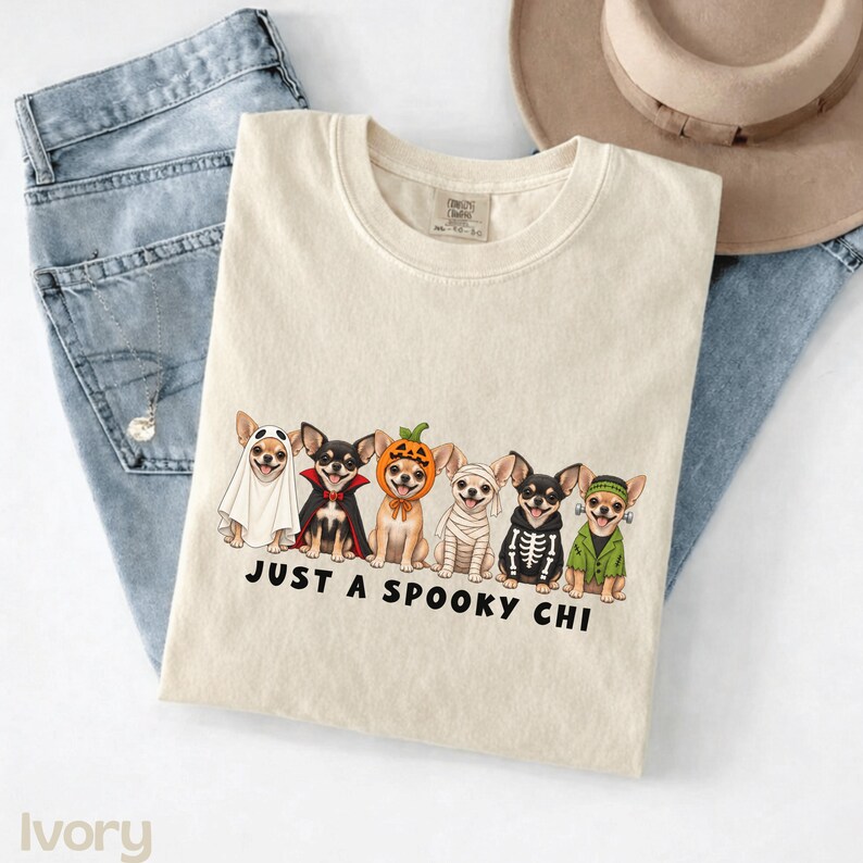 May include: An ivory crewneck t-shirt featuring a graphic of six Chihuahua dogs in various Halloween costumes, including a ghost, vampire, pumpkin, mummy, skeleton, and Frankenstein's monster, with the text JUST A SPOOKY CHI printed below.
