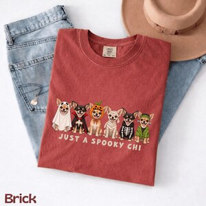 May include: A brick-colored t-shirt featuring a graphic of six Chihuahua dogs in various Halloween costumes, including a ghost, vampire, pumpkin, mummy, skeleton, and Frankenstein's monster, with the text "JUST A SPOOKY CHI" printed below.
