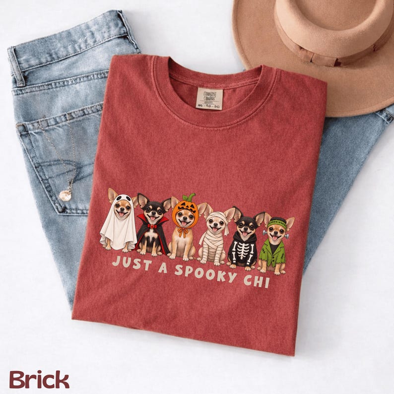 May include: A brick-colored t-shirt featuring a graphic of six Chihuahua dogs in various Halloween costumes, including a ghost, vampire, pumpkin, mummy, skeleton, and Frankenstein's monster, with the text "JUST A SPOOKY CHI" printed below.