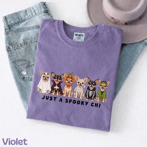 May include: A violet t-shirt featuring a graphic of six chihuahua dogs in various Halloween costumes, including a ghost, vampire, pumpkin, mummy, skeleton, and Frankenstein's monster, with the text "JUST A SPOOKY CHI" printed below.