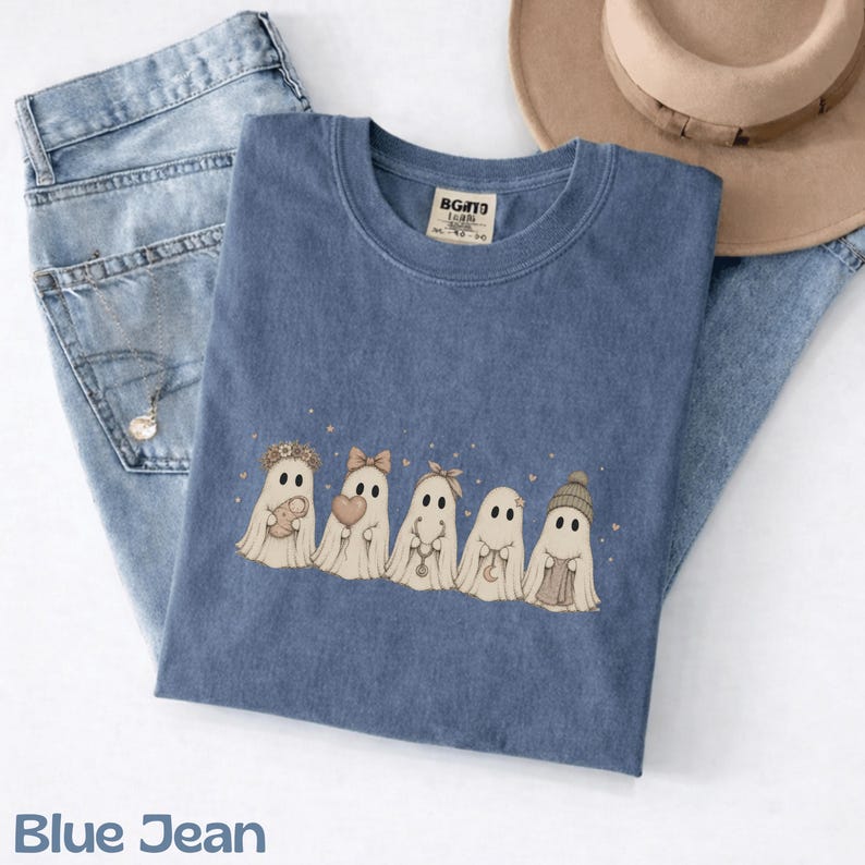 May include: A blue t-shirt featuring a graphic of five ghosts in a row. The ghosts are wearing accessories including a flower crown, a bow, a headband, and a knit hat, while holding items like a heart, a moon charm, and a small bundle.